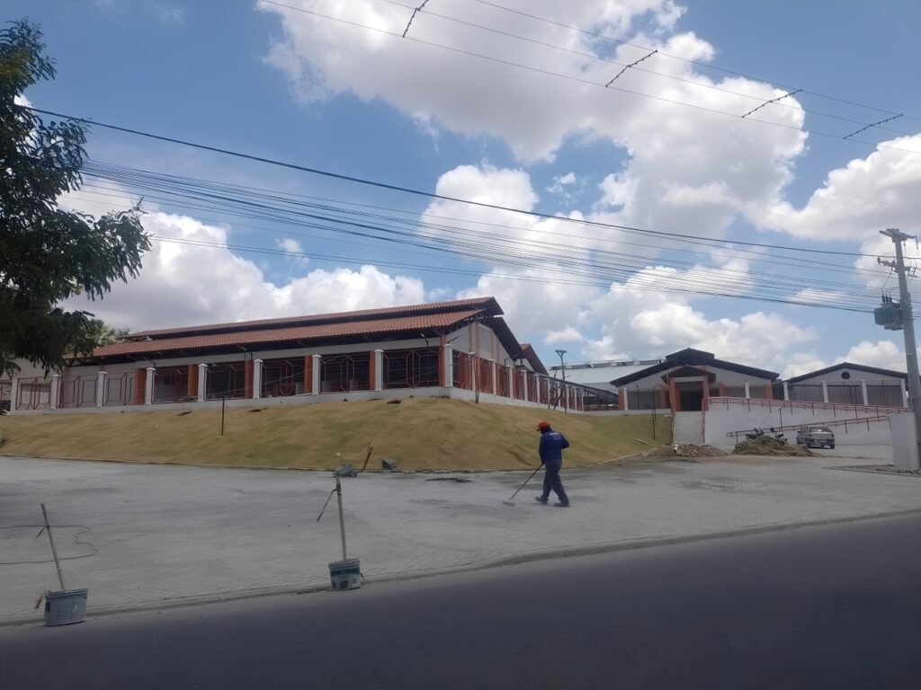 Obra da Escola Municipal Elma Camilo ao lado do terminal rodoviário de Gravatá com trabalhadores finalizando detalhes sob o sol do meio dia.