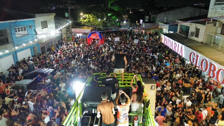 Fotografia colorida mostrando uma grande multidão de foliões lotando uma rua em Gravatá durante o desfile do Bloco DNA. Ao fundo, é possível ver estabelecimentos comerciais como a Padaria Globo e um portal inflável de cor azul e laranja da empresa Inovanet. A imagem captura o movimento intenso do pós-Carnaval na cidade.