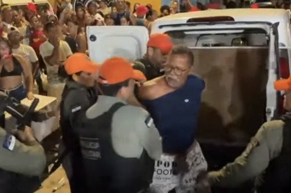 Policiais militares de Pernambuco abordam vendedor ambulante em frente a veículo Fiat Fiorino branco durante o bloco Ressaca em Caetés I, Abreu e Lima. Imagem registra momento da confusão que gerou investigação da Corregedoria da SDS por suspeita de agressão.