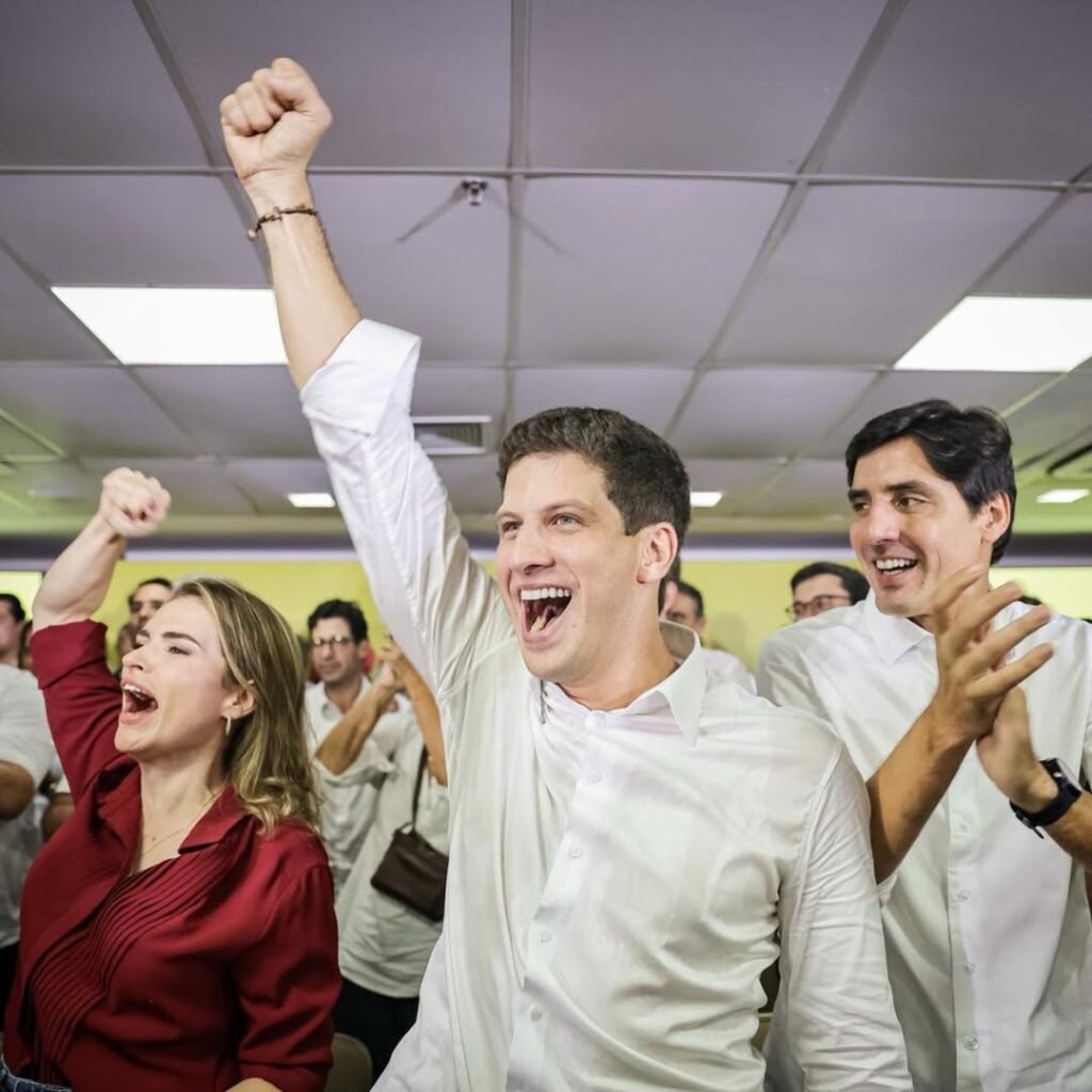 João Campos, Carlos Costa e Marília Arraes vibram de braços erguidos durante ato de lançamento da Frente Popular em Pernambuco. O registro mostra o prefeito do Recife (PSB), o pré-candidato a vice-governador e a pré-candidata ao Senado (PDT) em um momento de comemoração no palanque, sob luzes azuis e vermelhas, diante de uma militância entusiasmada e bandeiras do Brasil e de Pernambuco ao fundo.