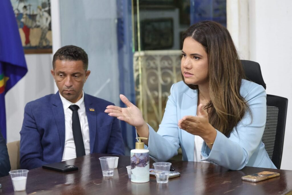 Prefeita de Olinda, Mirella Almeida, gesticula durante fala em ambiente institucional enquanto vice-prefeito aparece ao fundo em registro de arquivo