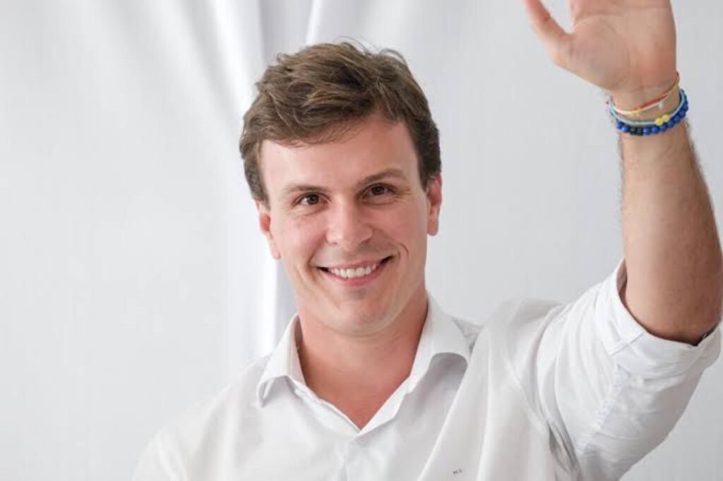 Fotografia de meio corpo de Miguel Coelho, sorrindo e acenando com a mão direita. Ele veste uma camisa social branca e está posicionado em frente a um fundo claro e desfocado. O pré-candidato ao Senado nas Eleições 2026 em Pernambuco transmite simpatia e proximidade em um registro de redes sociais, capturado durante um evento político.