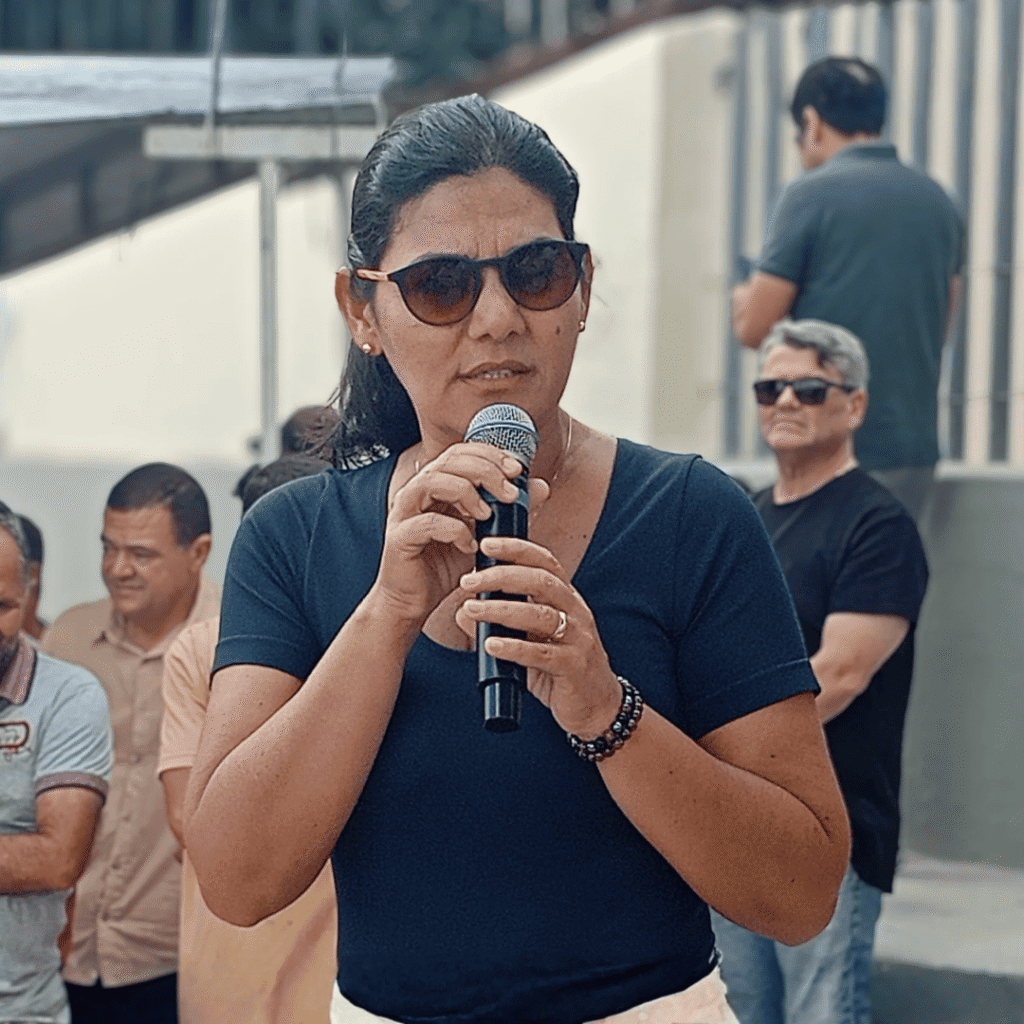 Viviane Facundes, secretária de Obras e primeira-dama de Gravatá, discursa com semblante sério e focado durante o ato de inauguração da urbanização da área frontal do Cemitério Santo Amaro. Ela está de pé, segurando o microfone com as duas mãos, transmitindo determinação enquanto fala contra a violência política e a misoginia. A imagem captura o momento exato de sua declaração de 'lugar de agressor é na cadeia', evidenciando sua postura de combate ao preconceito contra mulheres na política.