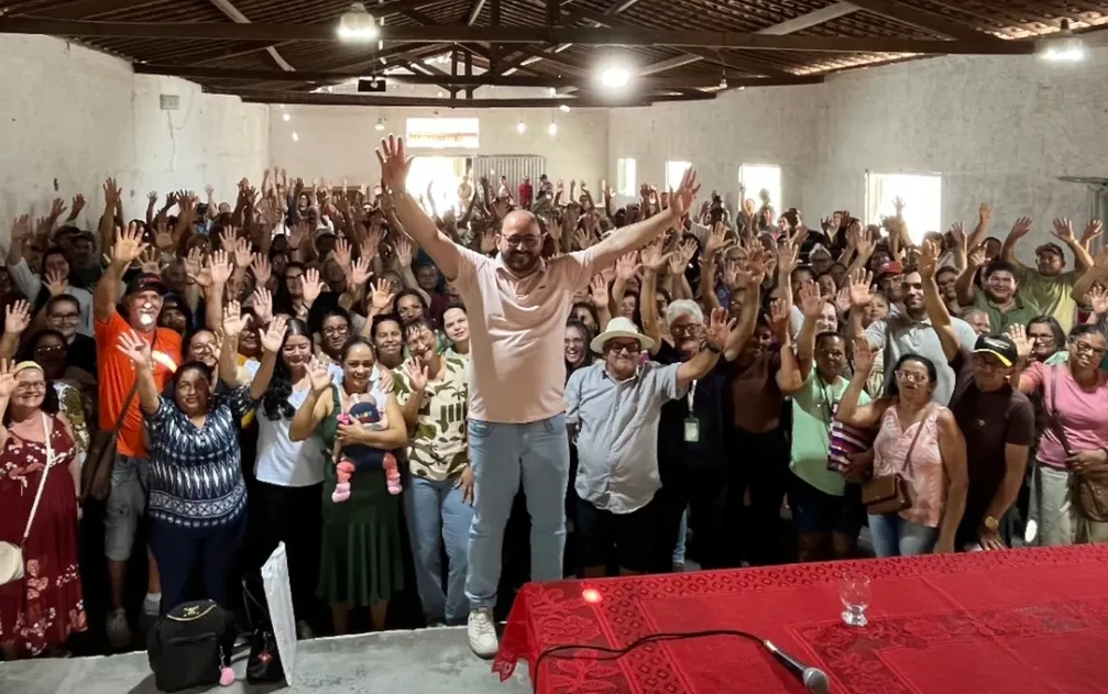 Fotografia tirada do palco de um auditório lotado durante a assembleia mensal do STR Gravatá. No centro da imagem, em pé e sorrindo com os braços erguidos em sinal de saudação, está Val Pessoa, coordenadora da nova gestão. Ela veste uma camisa polo rosa clara e calça jeans clara. Ao seu redor, centenas de agricultores e agricultoras associados levantam as mãos em resposta, demonstrando apoio e engajamento. O ambiente é um galpão amplo com estrutura de telhado de madeira à mostra e iluminação embutida. Em primeiro plano, uma mesa com toalha de renda vermelha e um microfone, indicando o local da presidência da reunião. A imagem transmite entusiasmo e comunhão.