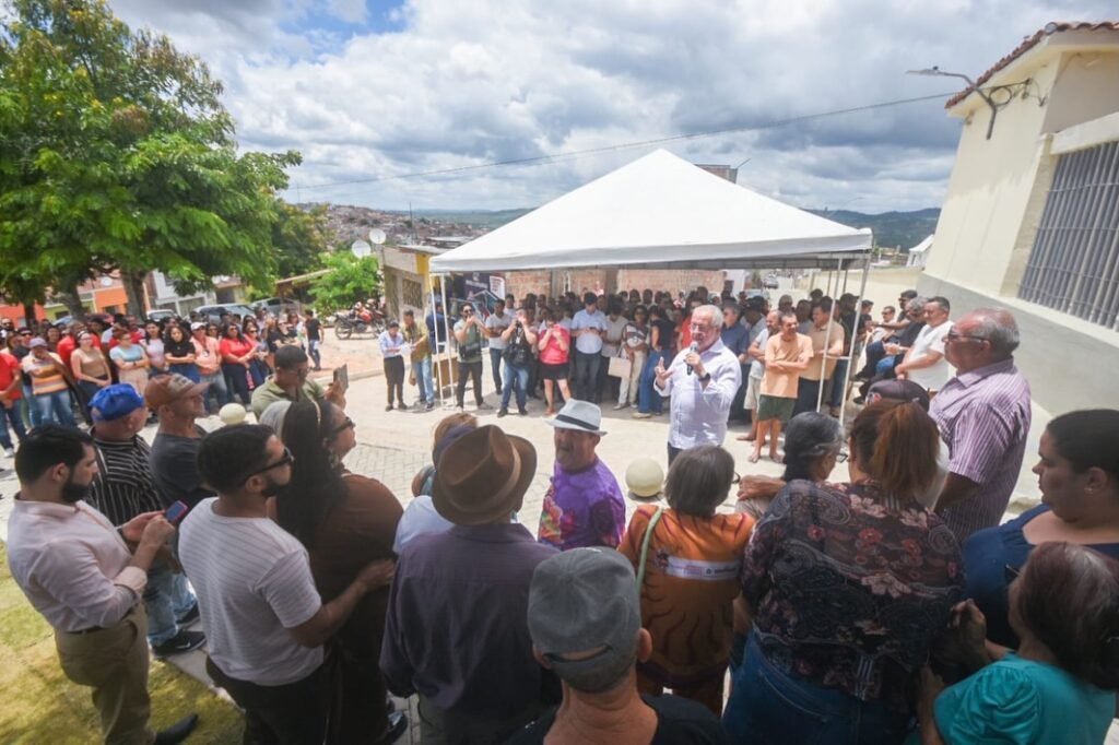 O prefeito de Gravatá, Joselito Gomes, discursa durante a inauguração da requalificação da área externa do Cemitério Santo Amaro. Ele aparece em destaque à frente de uma plateia de populares reunidos sob um céu limpo. Os moradores do Bairro do Cruzeiro acompanham atentos a entrega da obra, que conta com novos bancos de praça, gramado e iluminação moderna. A imagem, capturada por reprodução das redes sociais, reforça a presença da gestão municipal em eventos de entrega direta à população.