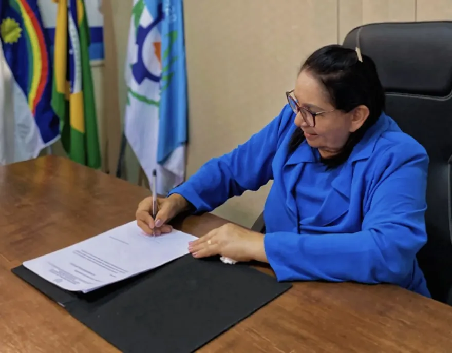 Arquivo fotográfico utilizado para ilustrar matéria sobre suspensão do concurso público de Catende após recomendação do MPPE por irregularidades. Na imagem, a prefeita Dona Graça aparece sentada em seu gabinete, assinando um documento em ambiente institucional, com semblante de riso discreto. 