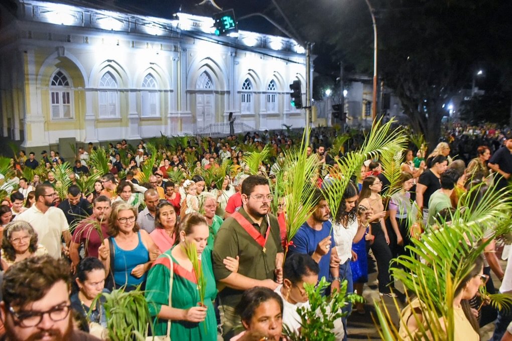 Uma multidão de centenas de fiéis caminha em procissão noturna pelas ruas de Gravatá, Pernambuco, segurando ramos de palmeiras verdes. No fundo, a imponente fachada amarela e iluminada do edifício histórico Paço Municipal está claramente visível.
