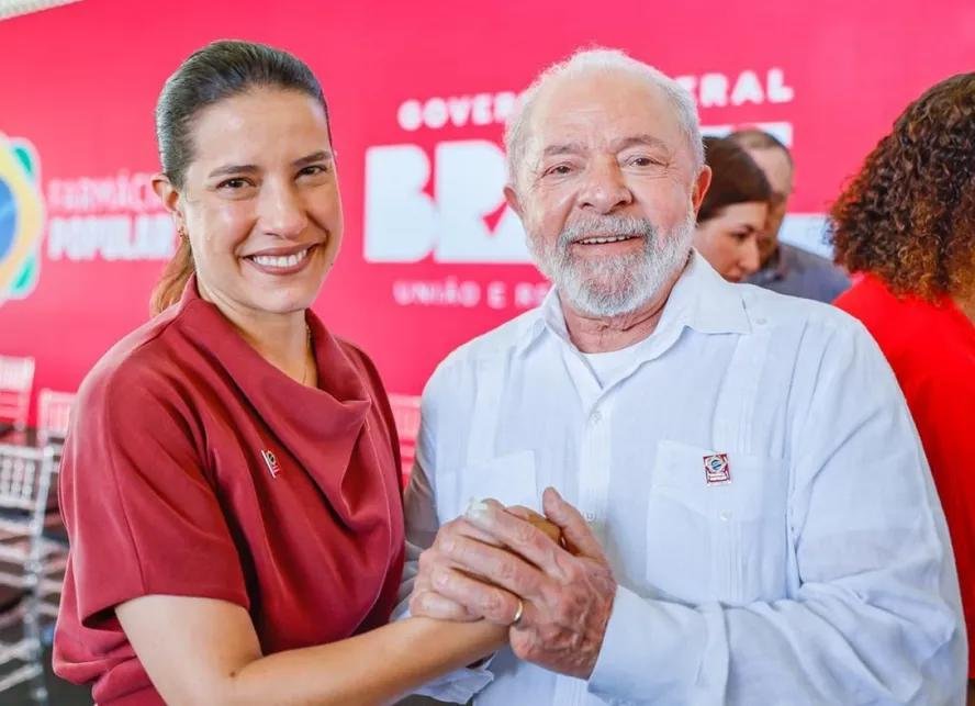 A governadora de Pernambuco, Raquel Lyra, vestindo uma blusa vermelha, aparece ao lado do presidente Lula em um palco durante evento institucional do Governo Federal. Eles estão sorridentes, gesticulando para o público. A composição da foto, assinada por Ricardo Stuckert, destaca o alinhamento institucional entre o Palácio do Campo das Princesas e o Planalto, com bandeiras oficiais ao fundo e iluminação frontal nítida.