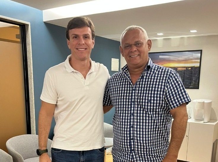 O ex-prefeito de Primavera, Rômulo César, e o pré-candidato ao Senado, Miguel Coelho, posam juntos e sorridentes para foto que oficializa o apoio político. Rômulo César (conhecido como 'Pão com Ovo') aparece à esquerda, vestindo uma camisa polo azul, e Miguel Coelho à direita, de camisa social branca. Ambos estão de pé, apertando as mãos e olhando para a câmera, simbolizando a aliança para as eleições de 2026 em Pernambuco. O fundo da imagem mostra um ambiente de escritório político com bandeiras de Pernambuco e do Brasil desfocadas