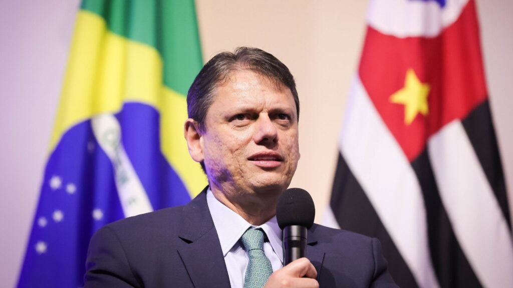 Governador Tarcísio de Freitas em pose oficial em ambiente institucional, vestindo terno escuro e gravata, com bandeiras ao fundo e expressão serena.