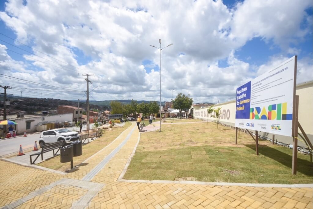 A área externa do Cemitério Santo Amaro, em Gravatá, após a reforma, exibe um amplo gramado verde bem cuidado, novos bancos de praça modernos e caminhos de paralelepípedo para acessibilidade. A foto, realizada sob um céu azul limpo e brilhante, destaca o novo urbanismo e a arborização, evidenciando o trabalho da Secretaria de Obras e Serviços Públicos no Bairro do Cruzeiro, sob a gestão do prefeito Joselito Gomes.