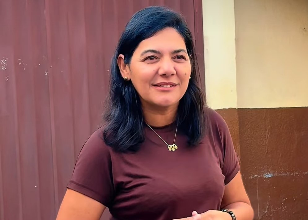 Fotografia colorida em plano médio de Viviane Facundes, mulher de cabelos escuros e sorriso suave, vestindo uma camiseta marrom-escura e colares dourados com pingentes. Ela está de pé, com as mãos entrelaçadas à frente, em um ambiente externo com fundo composto por uma porta de metal marrom texturizada e uma parede de concreto bege e marrom. A iluminação é natural e difusa.