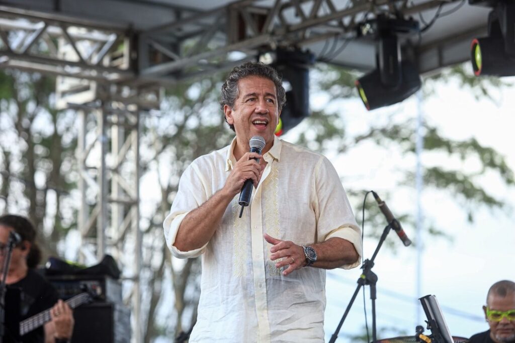 Fotografia em plano médio do cantor Altemar Dutra Jr. no palco do Alto do Cruzeiro, em Gravatá. Ele veste uma camisa social de linho em tom creme, com bordados sutis na frente, e um relógio no pulso esquerdo. Ele está cantando com um microfone na mão direita, voltado para a esquerda da imagem, com uma expressão emocionada. Ao fundo, desfocados, vê-se a estrutura metálica do palco e árvores sob a luz natural do dia.