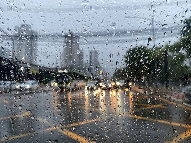 Vista embaçada de uma rua de cidade através de um vidro coberto por gotas de chuva. O fundo está desfocado, mostrando luzes de faróis de carros e prédios sob um céu cinzento. Imagem artística utilizada para ilustrar o alerta de chuvas fortes em Pernambuco emitido pela APAC.