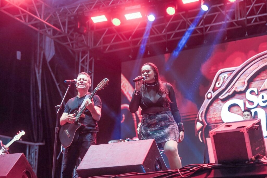 Fotografia de ângulo baixo do palco no Pátio de Eventos de Gravatá. À direita, uma cantora com cabelos longos, vestindo blusa preta transparente e saia brilhante, canta com o microfone na mão. À esquerda, um músico toca violão e canta em um pedestal. O palco está iluminado por fortes refletores vermelhos e brancos, com um grande painel de LED ao fundo exibindo a marca do evento Semana Santa Gravatá 2026.