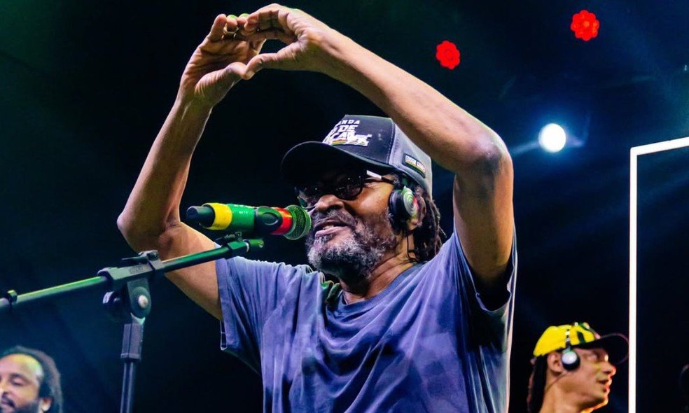O cantor Edson Gomes, um homem negro de barba grisalha e óculos, está no palco, visto de meio-corpo. Ele veste uma camiseta azul e um boné preto com detalhes em verde, amarelo e vermelho (cores do reggae), e fones de ouvido pretos. Com as duas mãos elevadas acima da cabeça, ele forma um símbolo de coração. Ele canta em um microfone colorido. O fundo é escuro, típico de um show, com luzes de palco vermelhas desfocadas. A imagem transmite alegria e conexão com o público.