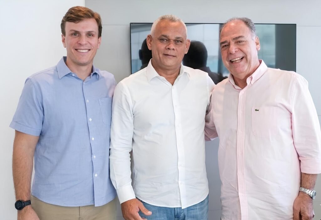 Fotografia de Miguel Coelho à esquerda, o ex-prefeito de Primavera Rômulo César (Pão com Ovo) ao centro e o ex-senador Fernando Bezerra Coelho à direita. Eles estão em pé, sorridentes, em um ambiente de escritório com iluminação clara.