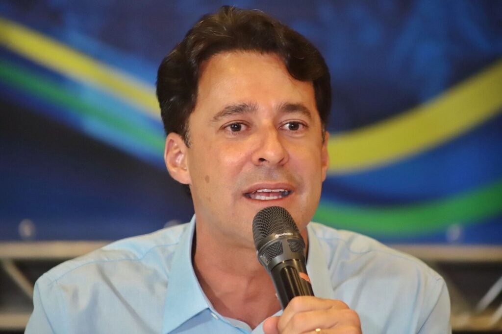 Um plano médio do político Anderson Ferreira, um homem de pele clara e cabelos escuros penteados, vestindo uma camisa de botão azul clara. Ele segura um microfone preto com a mão direita próximo à boca e está falando, com uma expressão tranquila e confiante. O fundo é desfocado, mostrando listras coloridas em azul, amarelo e verde. A imagem ilustra Anderson Ferreira em um momento de fala pública.