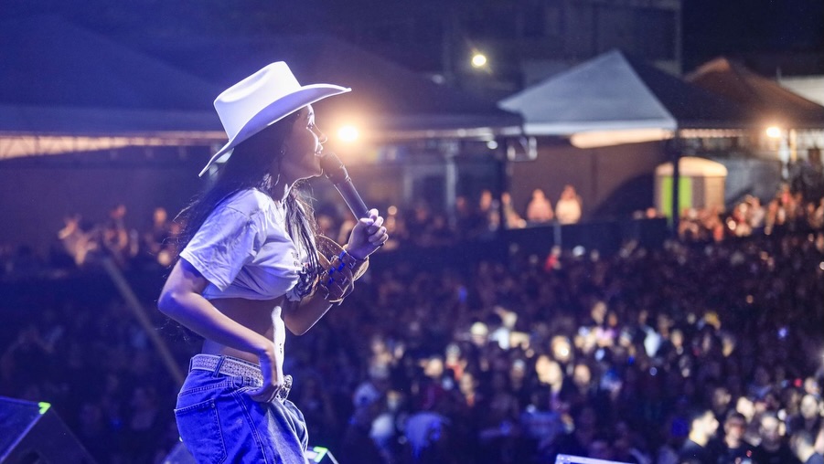 Fotografia de perfil da cantora Flay no palco do Pátio de Eventos de Gravatá. Ela veste um chapéu de cowboy branco, camiseta branca curta (cropped) e calça jeans, segurando um microfone próximo ao rosto. A cantora sorri enquanto olha para a plateia. Ao fundo, uma multidão desfocada sob luzes noturnas preenche o espaço do evento, com tendas brancas visíveis na linha do horizonte.