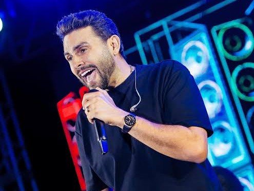 Uma fotografia vertical em plano médio do cantor Mano Walter no palco. Ele está posicionado do centro para a direita, segurando um microfone preto com a mão direita e sorrindo com a boca aberta, demonstrando animação. Mano Walter tem barba e usa uma camisa de manga curta preta básica e um relógio de pulso escuro com mostrador prateado no pulso esquerdo. O fundo é dinâmico, composto por luzes de palco LED azuis intensas e estruturas metálicas de treliça, com caixas de som empilhadas formando um padrão circular neon azul e ciano. Há uma luz branca brilhante no canto superior esquerdo e um vislumbre desfocado de outra pessoa na parte inferior esquerda. A iluminação geral é de show noturno, destacando o artista contra o fundo azul vibrante.