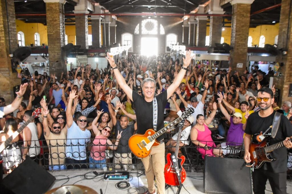 Fotografia em plano aberto capturada do palco do Mercado Cultural de Gravatá. No centro, o cantor Rodrigo Lins sorri com os braços abertos segurando uma guitarra Gibson Les Paul amarela. Ao seu lado, integrantes da banda Os Implacáveis. Ao fundo, uma multidão de pessoas de todas as idades lota o pavilhão histórico do mercado, com os braços levantados em sinal de festa. A iluminação natural entra pelos arcos do fundo do prédio.