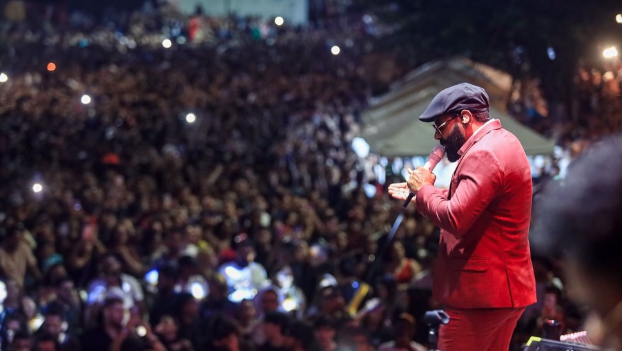 Fotografia em plano médio do cantor Pablo em cima de um palco, vestindo terno e calça vermelha e boina preta, cantando com microfone na mão. Ele está em pé no canto direito, voltado para o canto esquerdo, de perfil, com uma imensa multidão desfocada preenchendo o fundo, que está escuro. As pessoas na plateia seguram celulares com luzes e flashes acesos.