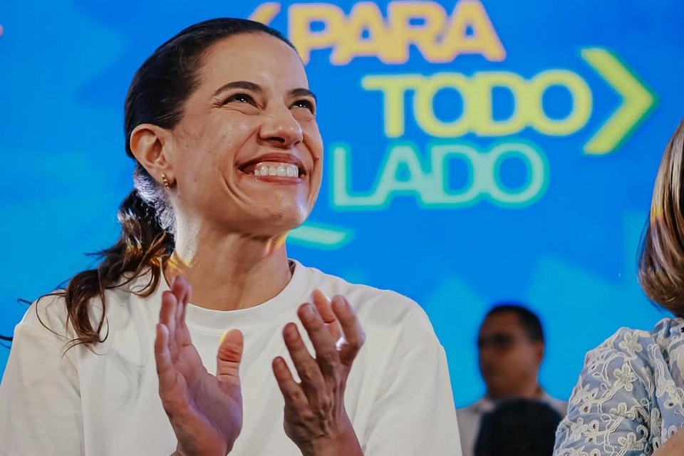 Governadora Raquel Lyra sorri e aplaude durante evento público, com painel azul ao fundo exibindo a frase Para todo lado