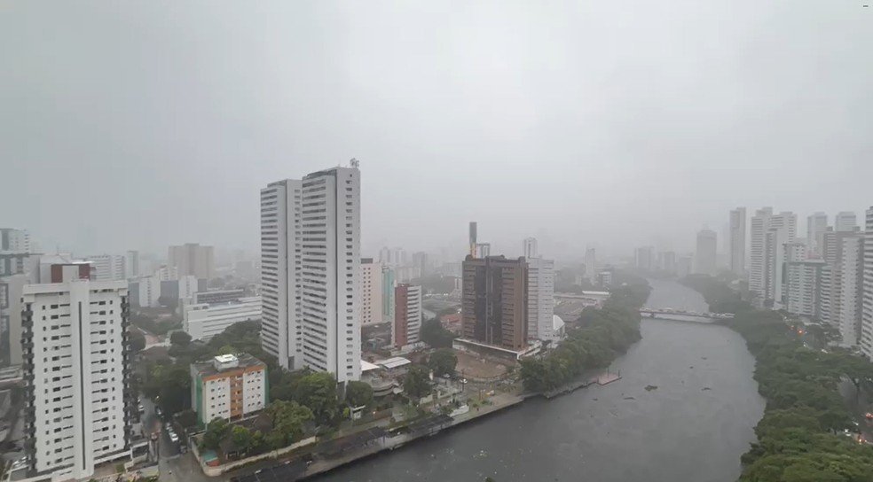 Vista aérea do Recife sob forte chuva e céu nublado. Edifícios altos na Zona Norte e o Rio Capibaribe com Recife Antigo desfocado ao fundo. Imagem tirada de um drone ilustra o alerta meteorológico na cidade.