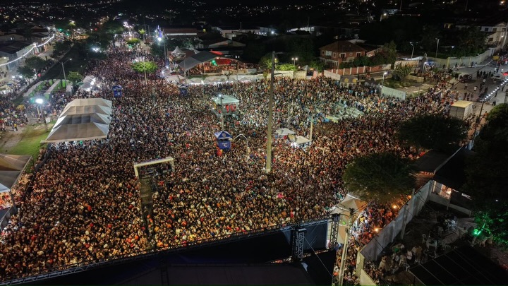 Fotografia aérea noturna em plano aberto do Pátio de Eventos Chucre Mussa Zarzar, em Gravatá. Uma multidão compacta de milhares de pessoas preenche todo o espaço da praça, cercada por tendas brancas nas laterais. Ao fundo, as luzes da cidade de Gravatá se estendem sob o céu escuro. A iluminação do evento cria um brilho dourado e vibrante sobre a massa de pessoas, destacando a magnitude do público durante a noite de sábado da Semana Santa 2026.