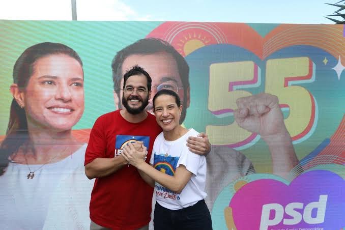 Plano médio de Túlio Gadêlha e Raquel Lyra, ambos sorrindo, abraçados e segurando a bandeira de Pernambuco. Túlio veste uma camisa vermelha com estampa da bandeira e Raquel uma camisa branca com a mesma estampa. Eles estão em pé diante de um grande banner publicitário. O banner tem um fundo colorido em tons de azul, verde, laranja e rosa, com a foto de Raquel Lyra ao fundo à esquerda e, à direita, o número "55" e o logotipo do partido "psd". A imagem simboliza a união e a aliança política entre os dois.