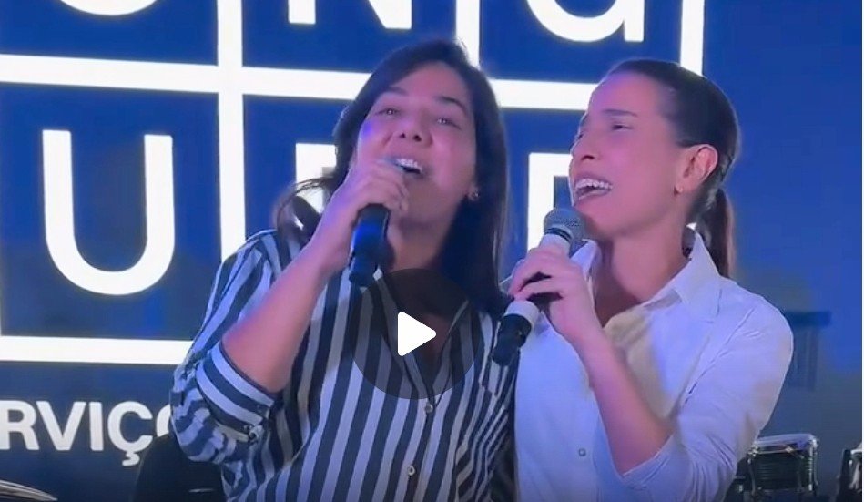 Viviane Facundes (à esquerda), com camisa listrada em preto e branco, e a governadora Raquel Lyra (à direita), com camisa branca social, cantam juntas no palco. Ambas seguram microfones próximos à boca e estão sorrindo, com olhos fechados ou semicerrados, demonstrando sintonia e alegria. O fundo é um painel de LED azul com letras brancas grandes, desfocadas.