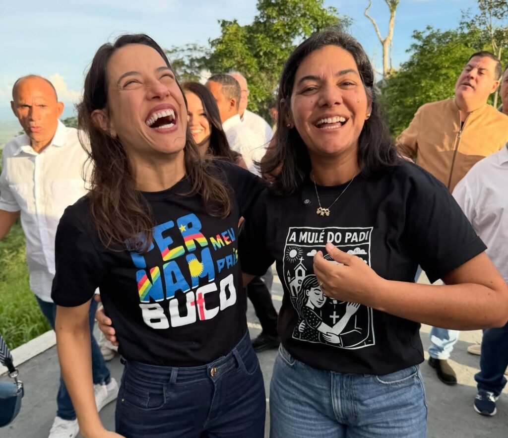 Fotografia de Raquel Lyra à esquerda e Viviane Facundes à direita. Ambas estão em um momento de descontração, rindo e abraçadas. Raquel veste uma camiseta preta com a estampa "Pernambuco Meu País" e Viviane usa uma camiseta preta com a estampa "A Mulé do Padi". Elas estão ao ar livre durante uma agenda oficial em Taquaritinga do Norte.