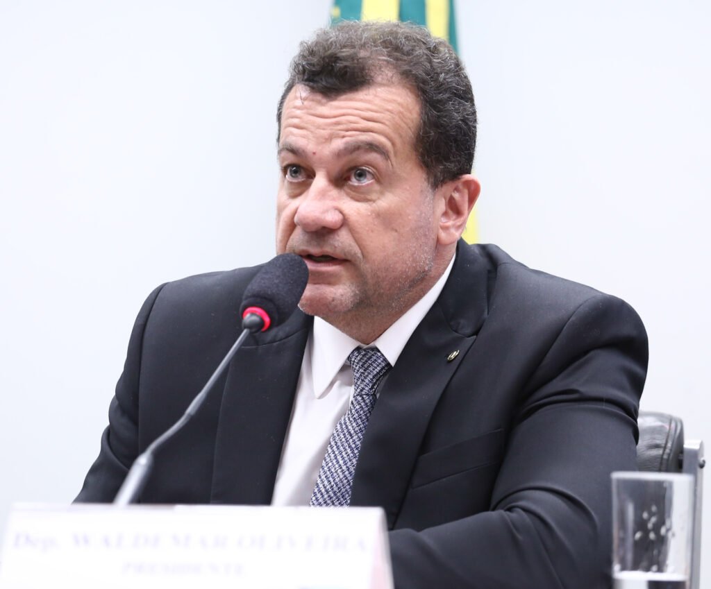 Fotografia de plano médio do deputado federal Waldemar Oliveira em ambiente institucional. Ele está falando ao microfone, veste terno preto, camisa branca e gravata cinza com padronagem discreta. O parlamentar tem expressão séria e focada. Ao fundo, detalhes de uma bandeira e, em primeiro plano, parte de uma placa de identificação na mesa da Câmara dos Deputados.