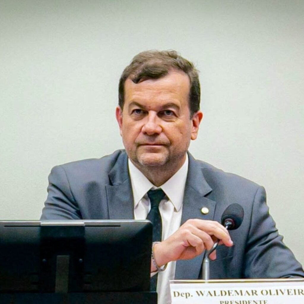 Deputado federal Valdemar Oliveira (Avante-PE), líder da bancada, de terno cinza, segurando microfone em mesa de comissão na Câmara dos Deputados em Brasília. A foto mostra sua placa de identificação com o título de "Presidente".