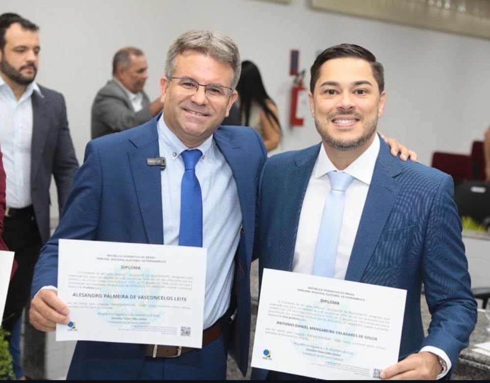 Uma fotografia horizontal em plano médio de dois homens sorrindo, em pé e lado a lado, em um ambiente interno bem iluminado. À esquerda está Alessandro Palmeira (Sandrinho), prefeito, usando óculos de grau, um terno azul-marinho, camisa social azul-clara e uma gravata azul-royal vibrante. Ele segura com a mão esquerda um diploma oficial de papel branco com o logotipo do TRE-PE e o nome "ALESSANDRO PALMEIRA DE VASCONCELOS LEITE" visível. Sua mão direita está sobre o ombro do homem ao lado. À direita está Daniel Valadares, vice-prefeito, vestindo um terno azul-petróleo textured, camisa social branca e uma gravata azul-clara listrada. Ele segura com a mão direita seu próprio diploma oficial idêntico, onde se lê "ANTONIO DANIEL MANGABEIRA VALADARES DE SOUZA". Ambos seguram os documentos na altura do peito e olham diretamente para a câmera, sorrindo. O fundo está desfocado, mas mostra outras pessoas vestindo trajes formais e uma parede com uma sinalização vermelha e branca vertical (possivelmente um extintor ou sinal de emergência). A iluminação é difusa, realçando os detalhes dos trajes e dos documentos.