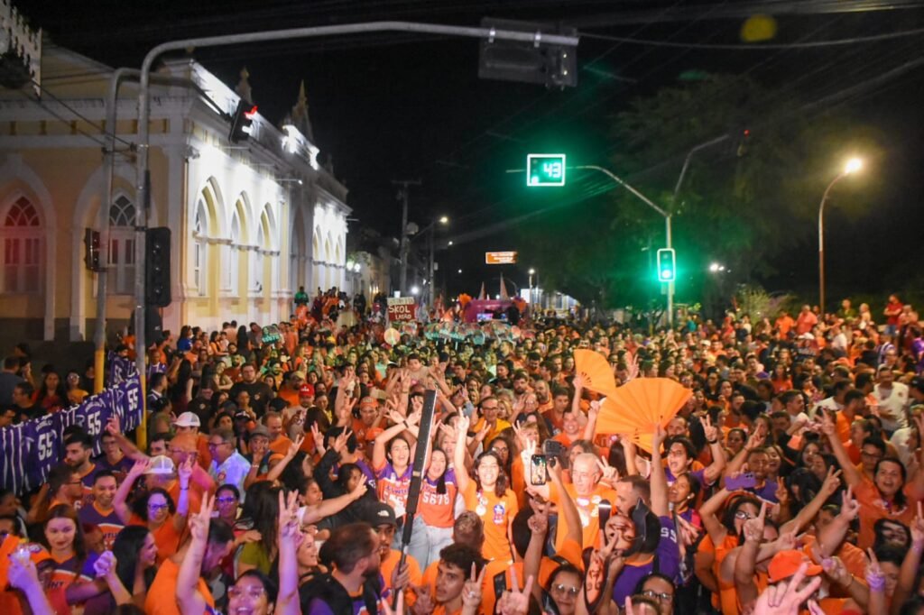 Multidão vestindo roupas laranjas e roxas reunida à noite na rua durante evento político de Viviane Facundes em Gravatá, em frente a um prédio histórico iluminado.