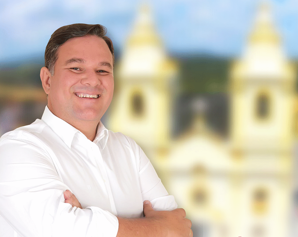 Ex-vereador Sérgio do Sindicato sorrindo de braços cruzados com camisa social branca, tendo ao fundo a Igreja Matriz de Chã Grande desfocada.