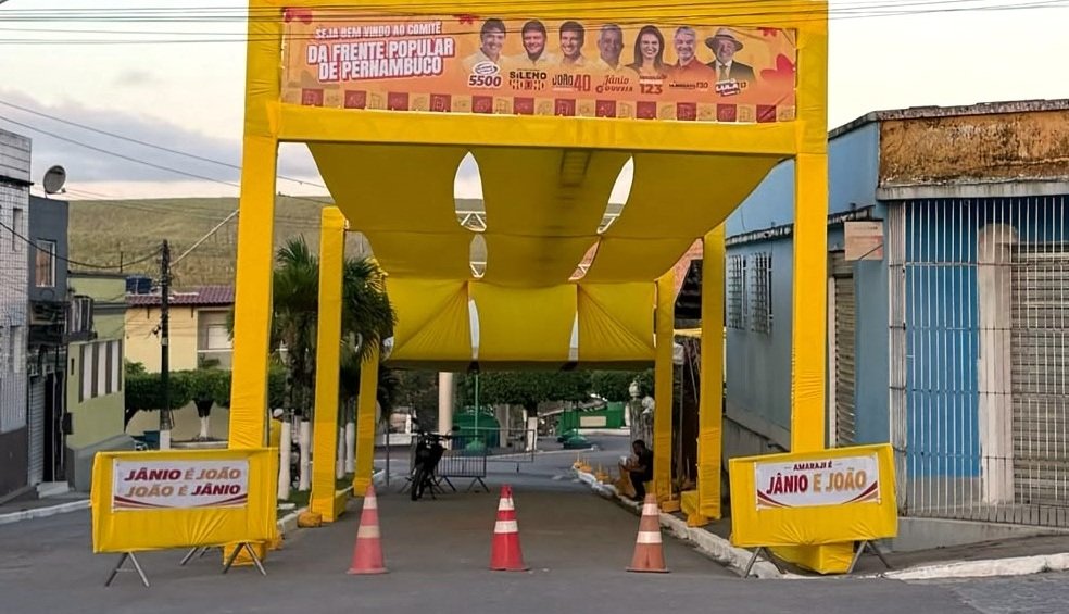 Pórtico amarelo de grande porte instalado sobre rua pública no Centro de Amaraji, contendo banner eleitoral da Frente Popular de Pernambuco e cavaletes bloqueando o trânsito.