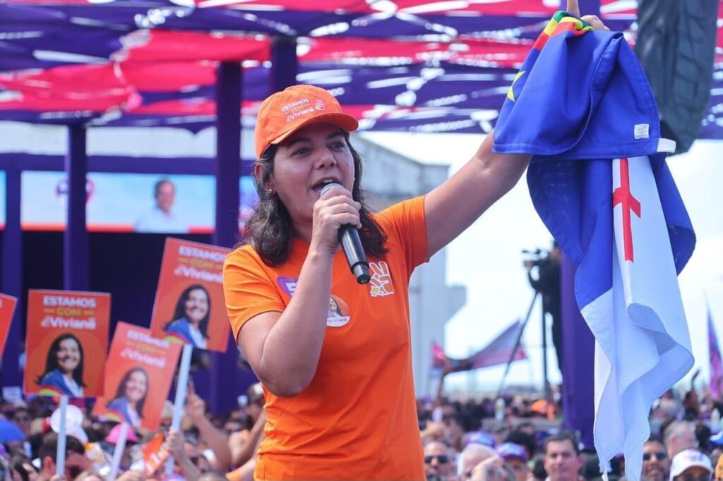 Viviane Facundes, de boné e camisa laranja, discursa com microfone e ergue a bandeira de Pernambuco na convenção do PSD no Recife.