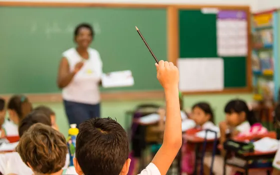 Estudante erguendo a mão com um lápis em primeiro plano durante aula em escola municipal de Gravatá, com professora ao fundo diante do quadro escolar.