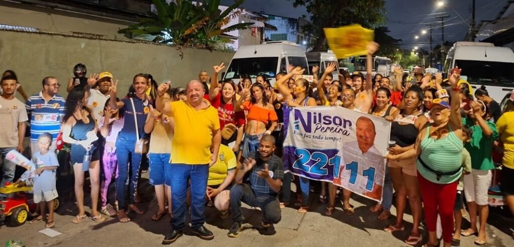 Nilson Pereira ao centro, vestindo camisa amarela e calça jeans, sorrindo e fazendo gesto com a mão, cercado por apoiadores reunidos à noite no bairro da Mustardinha, Recife, segurando uma faixa de campanha e uma bandeira amarela.