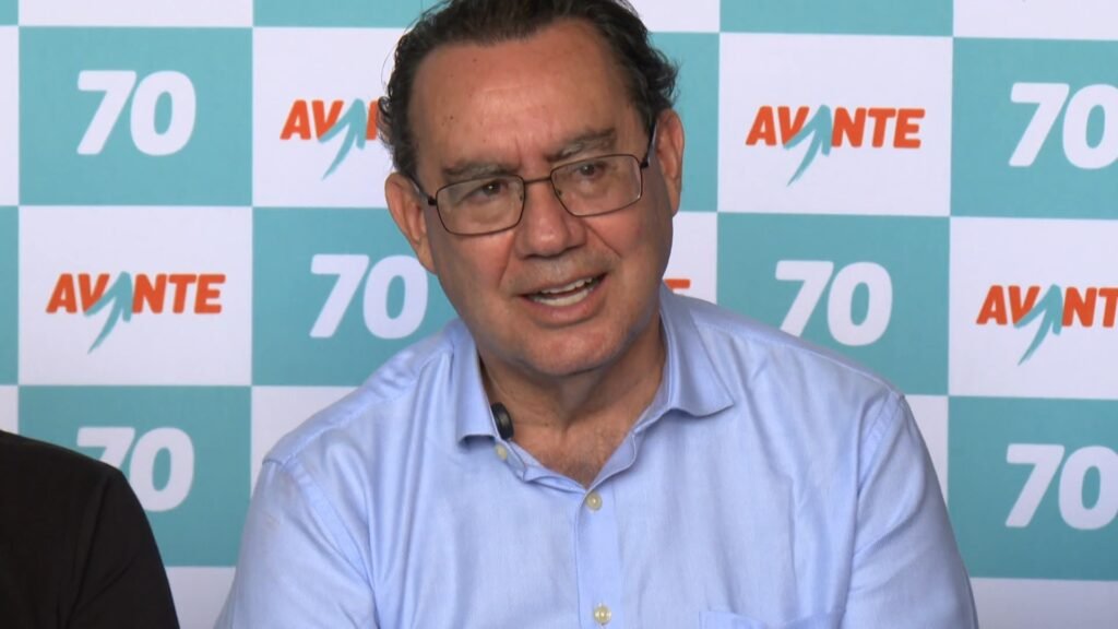 Augusto Cury, candidato à Presidência da República pelo Avante, usando óculos e camisa azul clara diante de um painel com o logotipo do partido Avante.