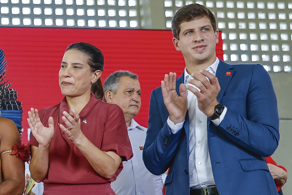João Campos e Raquel Lyra aplaudem de pé durante evento oficial, com homem ao fundo e presença de lideranças indígenas à esquerda na imagem.