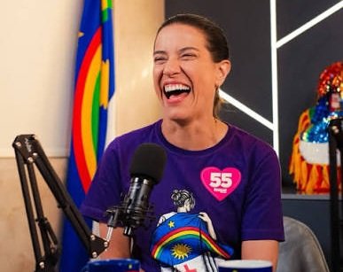 A governadora de Pernambuco, Raquel Lyra, sorri durante entrevista em estúdio de podcast usando camisa roxa com o número 55 do PSD e estampa da bandeira de Pernambuco.