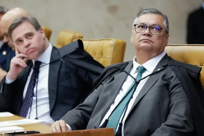 Uma fotografia em plano médio mostra dois ministros do Supremo Tribunal Federal (STF) sentados lado a lado em uma sessão plenária. Em primeiro plano e à direita, o ministro Flávio Dino, de óculos e terno escuro com gravata verde e a toga preta do STF, olha atentamente para a frente. Ao seu fundo e à esquerda, o ministro André Mendonça, também de terno escuro e toga, observa a cena com a mão direita apoiada no queixo. Eles estão sentados em poltronas de couro amarelo, em uma sala com painéis de mármore bege ao fundo.