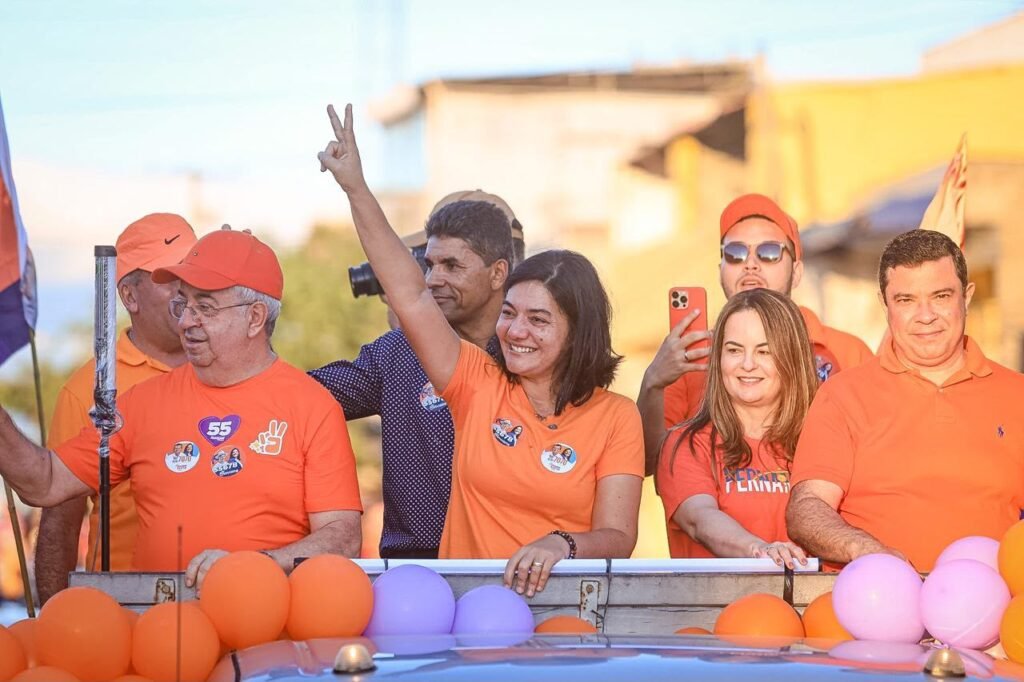 Uma fotografia em close-up de Viviane Facundes sorrindo e fazendo o sinal de vitória com a mão direita em cima de um veículo. Ela está cercada por apoiadores e pelo prefeito Joselito Gomes, todos vestindo camisetas laranjas de campanha. Ao redor, há muitos balões laranjas. O fundo é uma rua em Gravatá com construções baixas e céu azul.