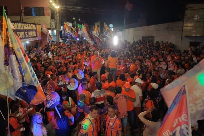 Fotografia noturna em ângulo elevado mostrando uma multidão lotando uma rua iluminada durante ato político de campanha. Apoiadores vestem roupas laranjas, seguram bandeiras e placas com o número 55678 e o nome de Viviane Facundes. Ao centro, lideranças discursam com microfone.