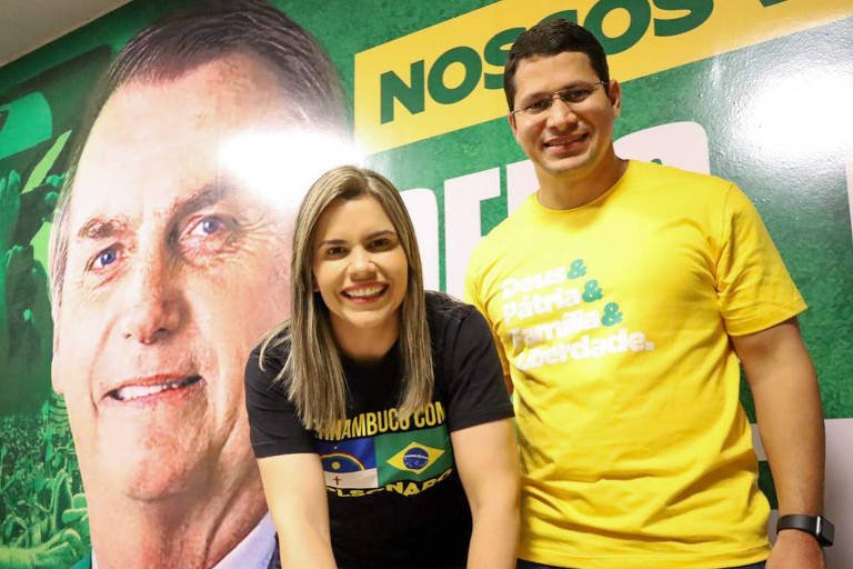 Clarissa Tércio e Júnior Tércio sorriem lado a lado em foto oficial. Ela veste camisa preta com a bandeira de Pernambuco e do Brasil, e ele veste camisa amarela com termos estampados. Ao fundo, há um painel com a foto do ex-presidente Jair Bolsonaro.