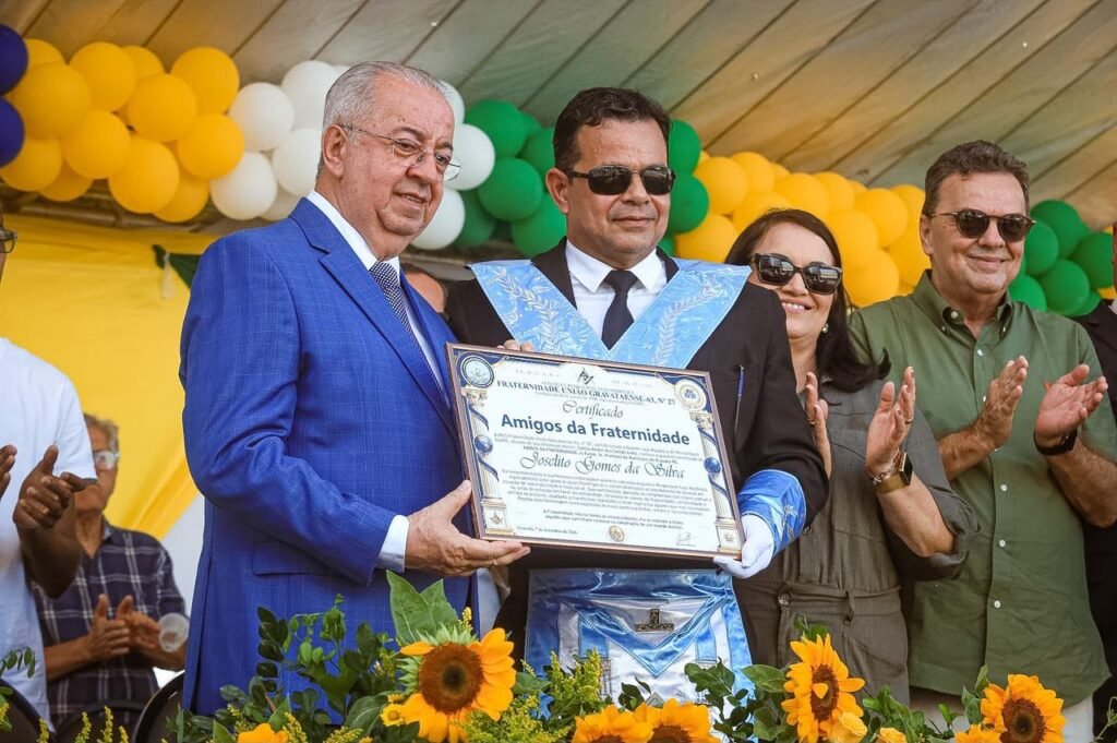 "Prefeito de Gravatá, Joselito Gomes da Silva (esq., terno azul), recebe o certificado 'Amigos da Fraternidade' de um representante da Loja Maçônica Fraternidade União Gravataense-63. O homenageado sorri e segura o documento emoldurado. O maçom veste paramento ritualístico azul e luvas brancas. Ao fundo, balões amarelos e verdes e girassóis na decoração. Gravatá, Pernambuco."