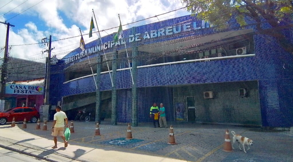 Fachada do prédio da Prefeitura Municipal de Abreu e Lima em tom azul, com pedestres e cones de trânsito na calçada, ilustrando a recomendação do MPPE para pagamento de diferenças salariais a professores.